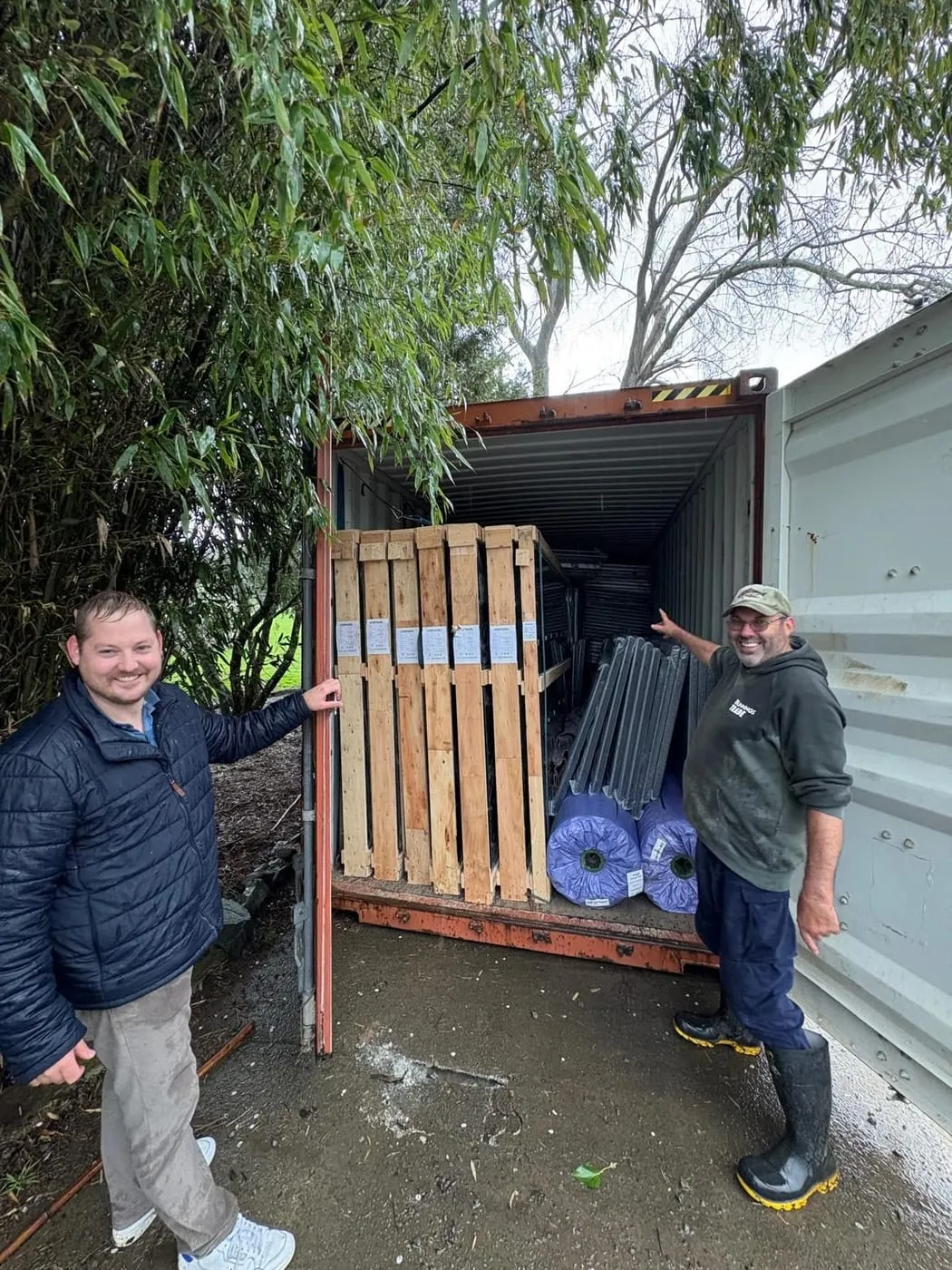Two people smiling beside a shipping container packed with crated court frames and turf rolls