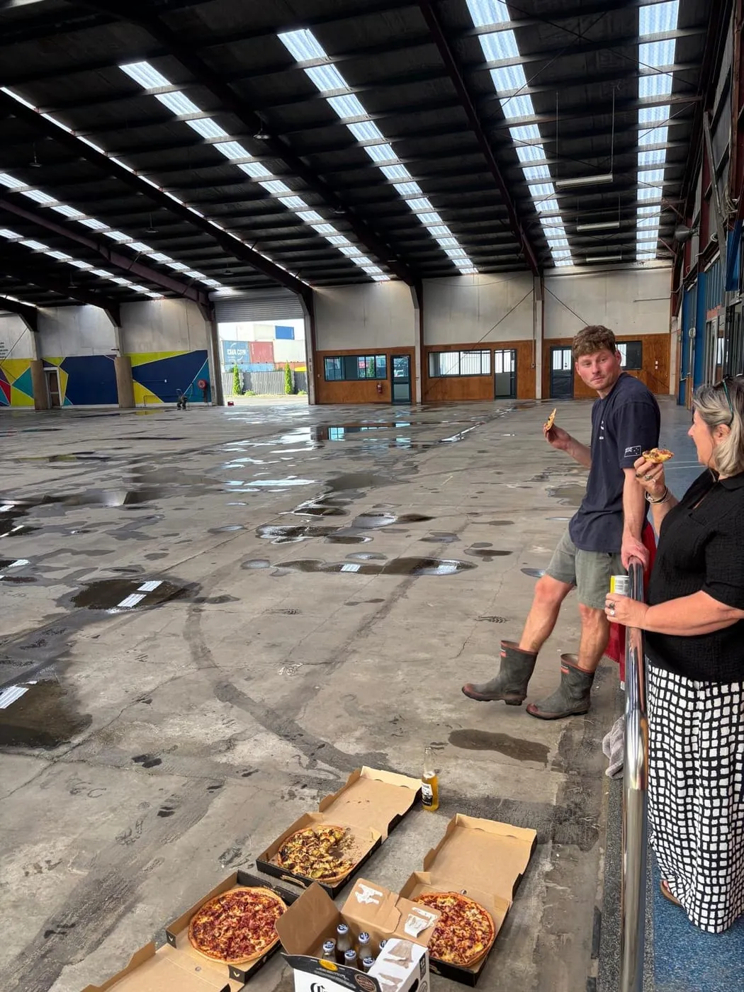 Two people eating pizza in the empty warehouse, puddles on the concrete