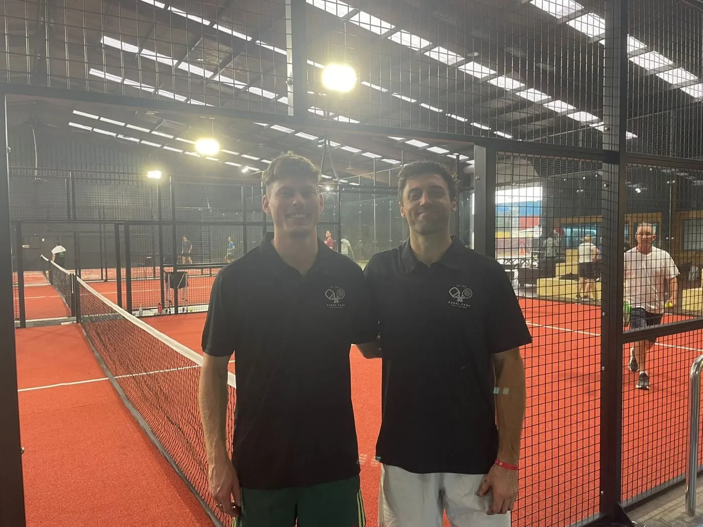 Two Padel Park staff in black polos smiling on court, players behind them